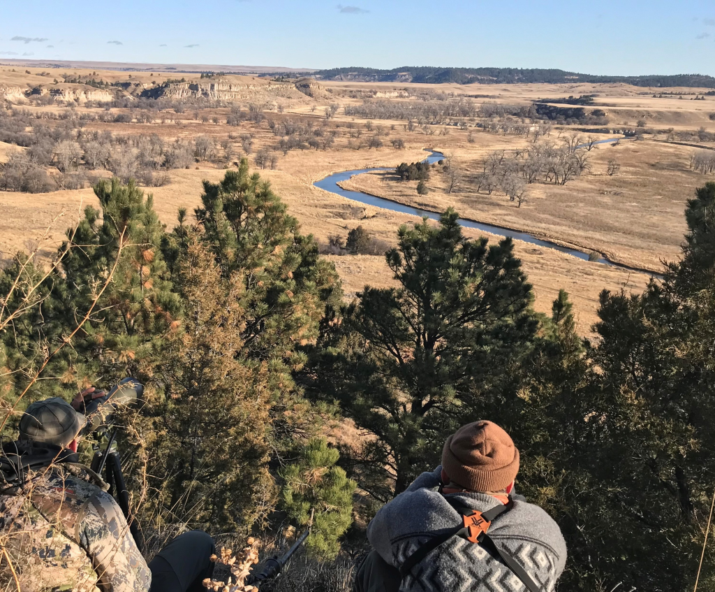 Two hunters glassing from a wooded hillside across open prairie and river-bottom country.