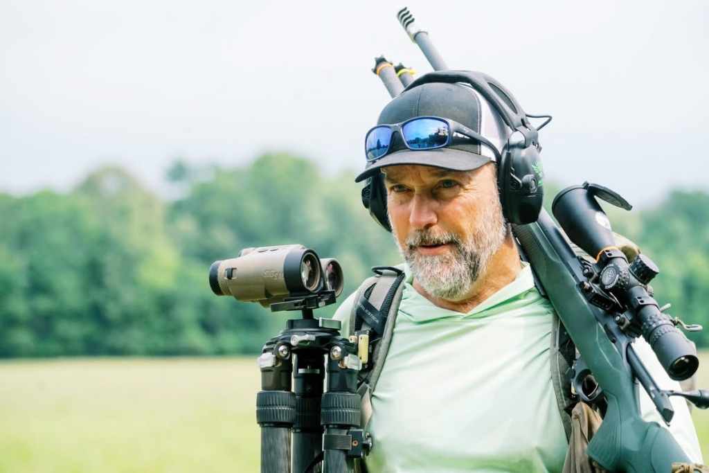Precision rifle competitor walking between NRL Hunter stages with a rifle, tripod and binoculars.