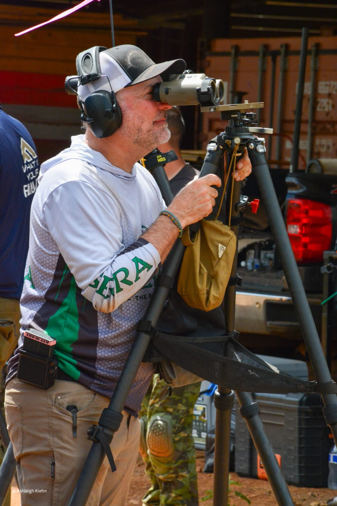 Precision rifle competitor watching other shooters through binoculars mounted on a tripod during a PRS match.